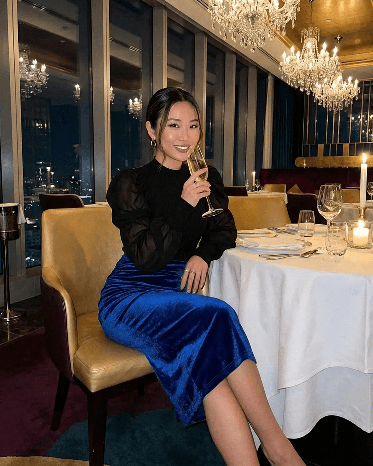 Woman in Azure Midi Skirt by SLIMONA enjoys a drink at an elegant restaurant setting.