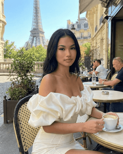 Celeste Mini Dress by SLIMONA, featuring off-the-shoulder design and ruffle trim, model enjoying coffee in Paris.