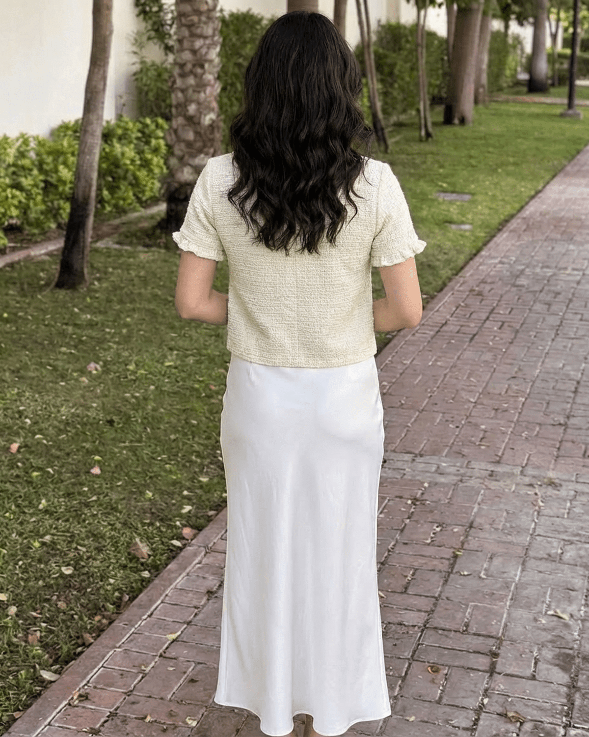 Back view of a woman wearing the Elara Cardigan by SLIMONA with a white skirt in a garden setting.