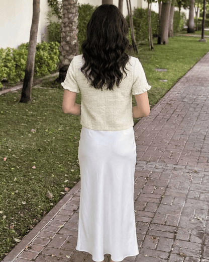 Back view of a woman wearing the Elara Cardigan by SLIMONA with a white skirt in a garden setting.