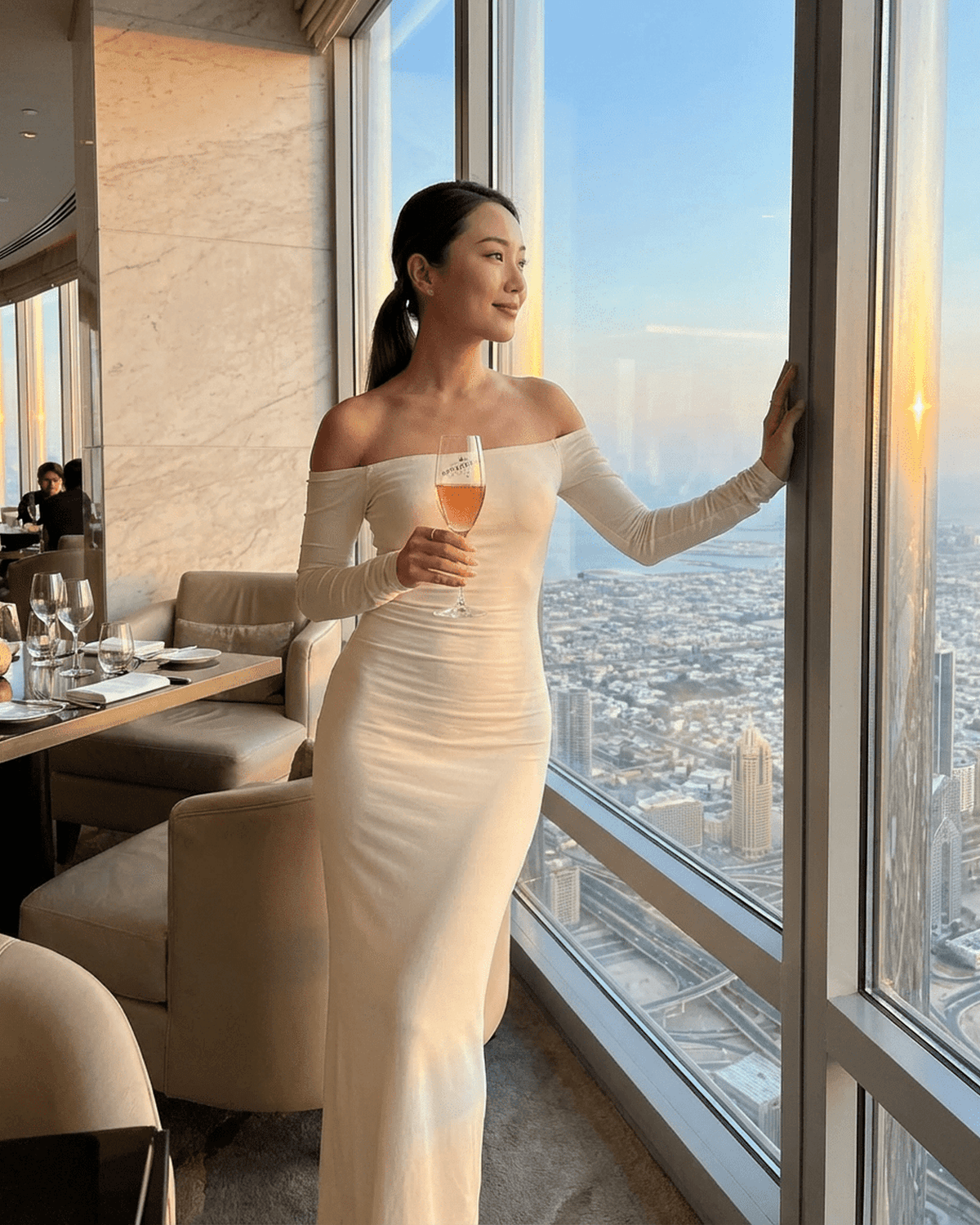 Elegant woman in Ivory Maxi Dress by SLIMONA, off-shoulder design, enjoying a sunset view with a drink.