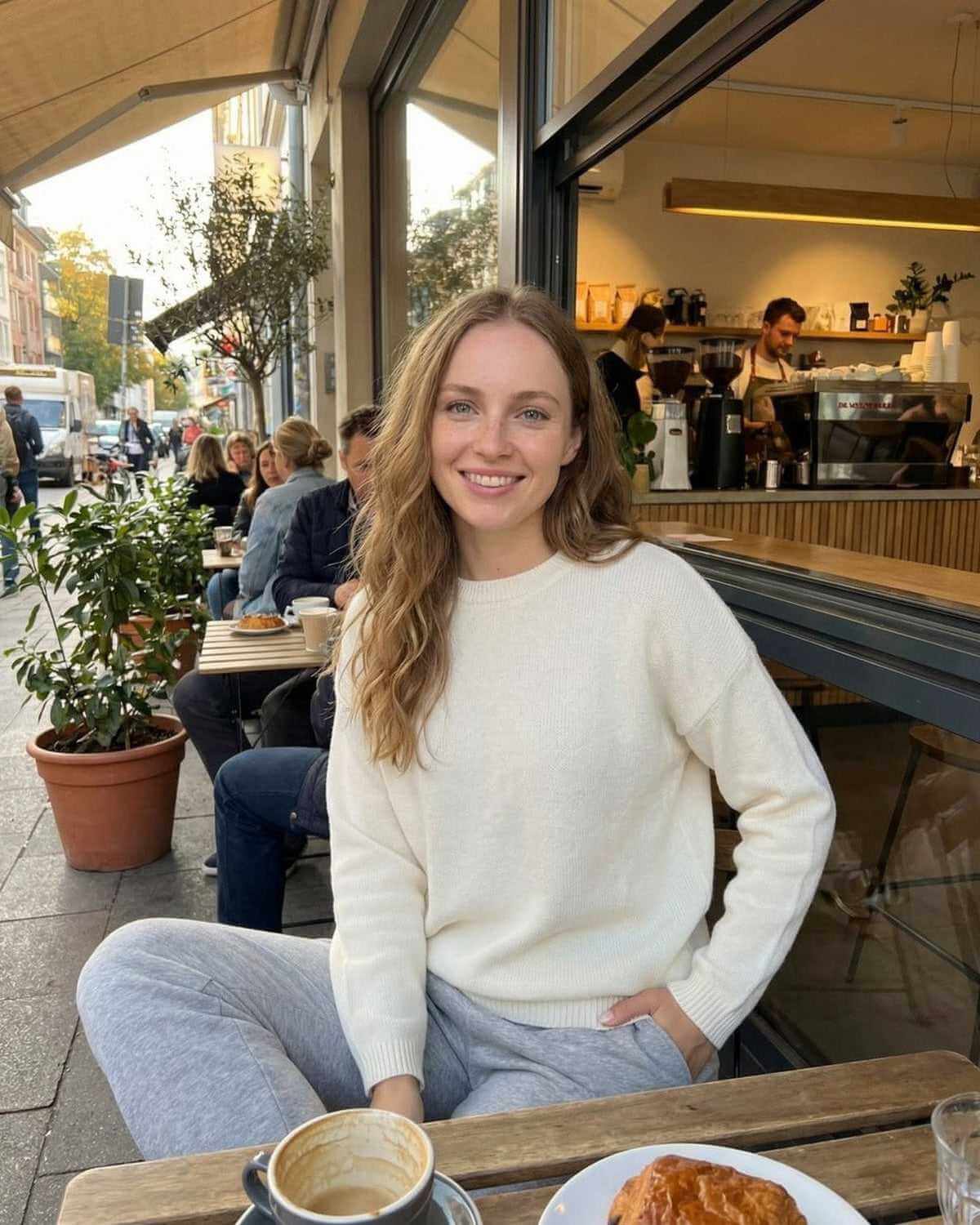 Woman wearing Alba Pullover by SLIMONA, enjoying coffee outdoors in casual setting.