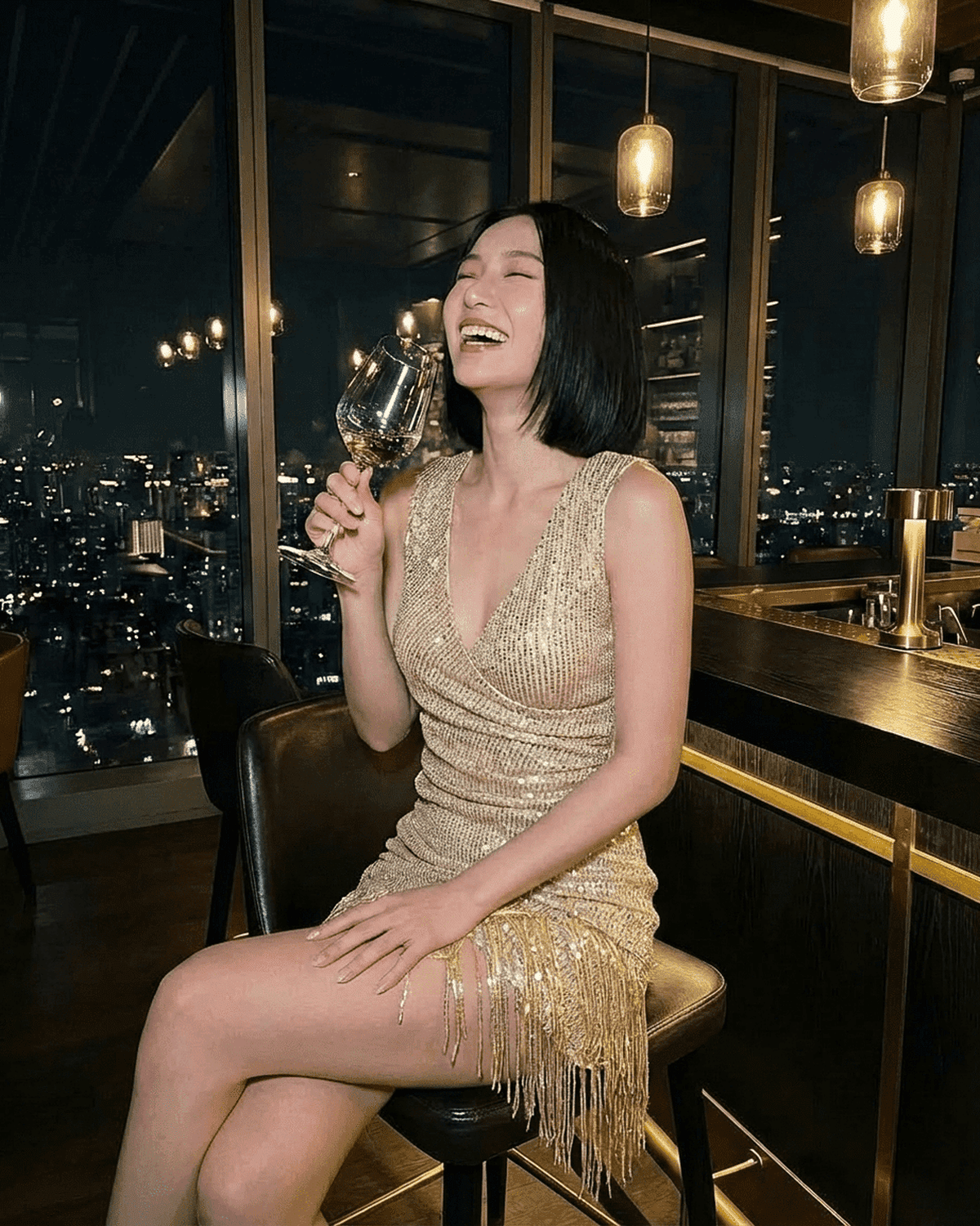 Smiling woman in Aurora Mini Dress by SLIMONA, a gold sequin V-neck midi with fringe hem, enjoying a drink.