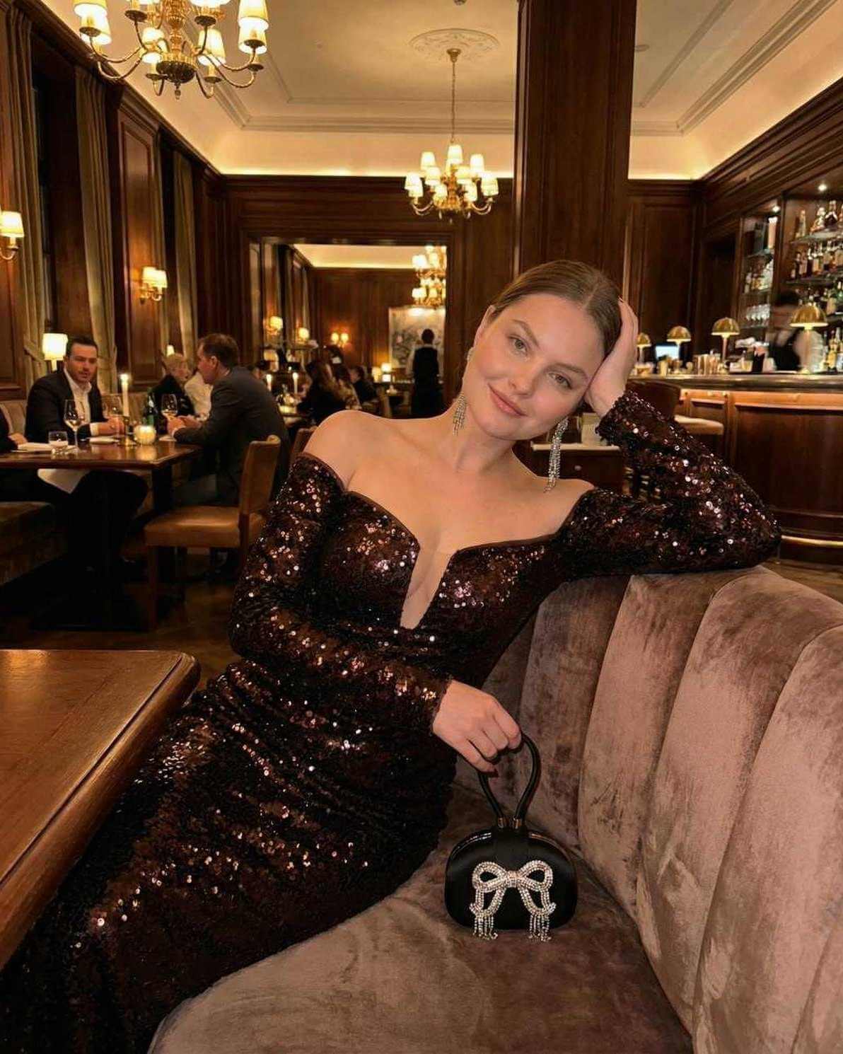 Celeste Maxi Dress by Lauven, an elegant sequined off-shoulder evening gown in deep brown worn in a sophisticated dining setting.