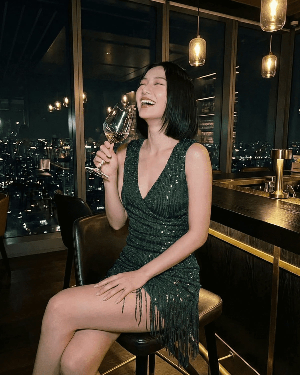 Woman enjoying a drink in an Emerald Mini Dress by SLIMONA, featuring green sequins and fringe hem.