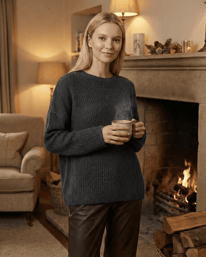 Woman wearing the Vega Sweater by SLIMONA, enjoying a cozy moment by the fireplace with a warm drink.