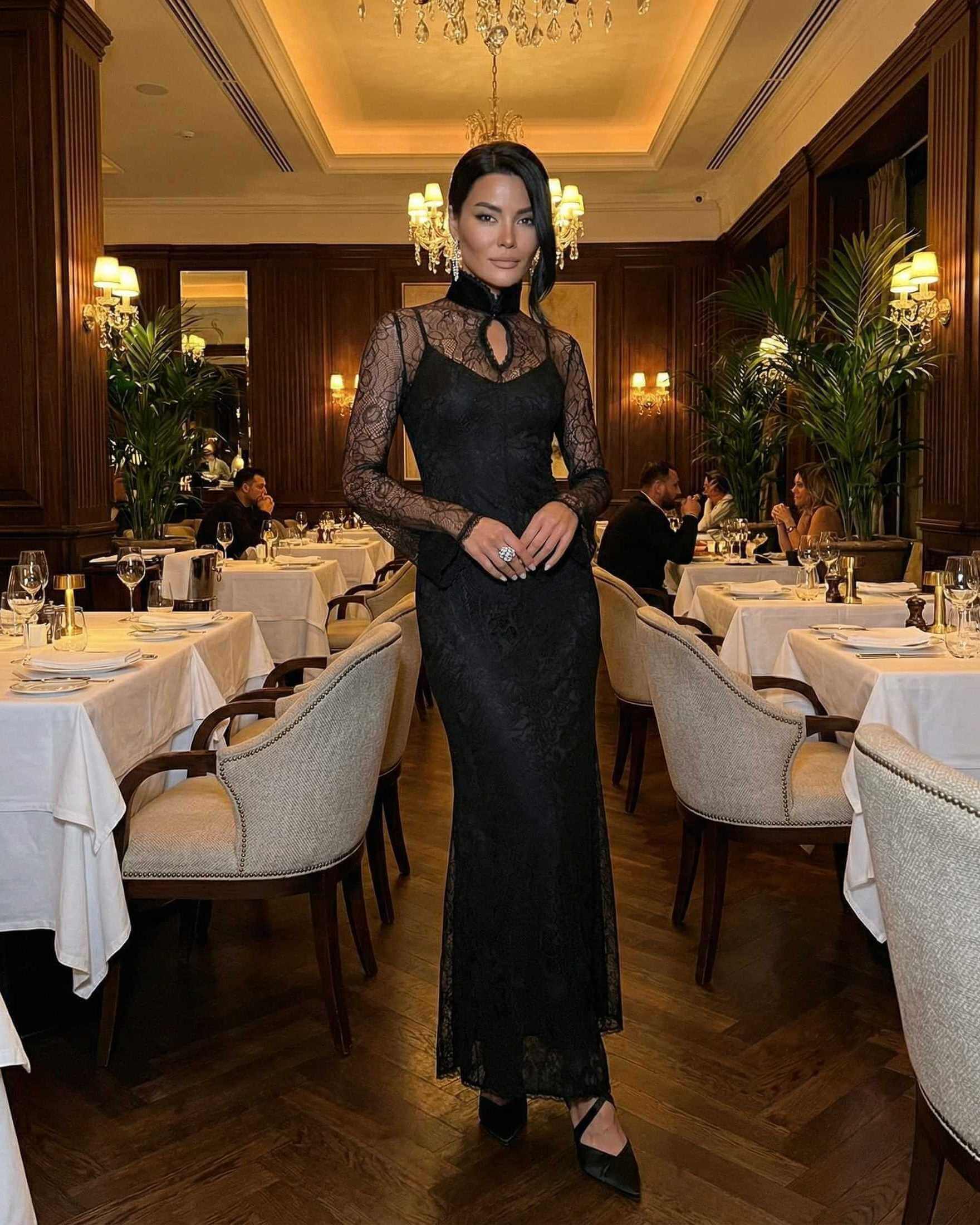 Woman wearing Noctavia Maxi Dress by Lauven, black lace long sleeve bodycon dress with stand collar in elegant restaurant.