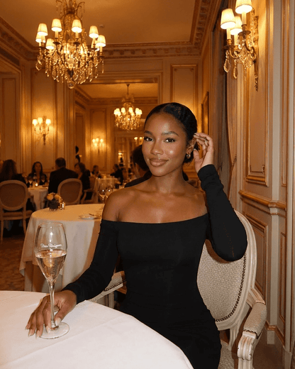 Elegant woman in Nocturne Maxi Dress by SLIMONA, sitting at a luxurious restaurant table with a glass of champagne.