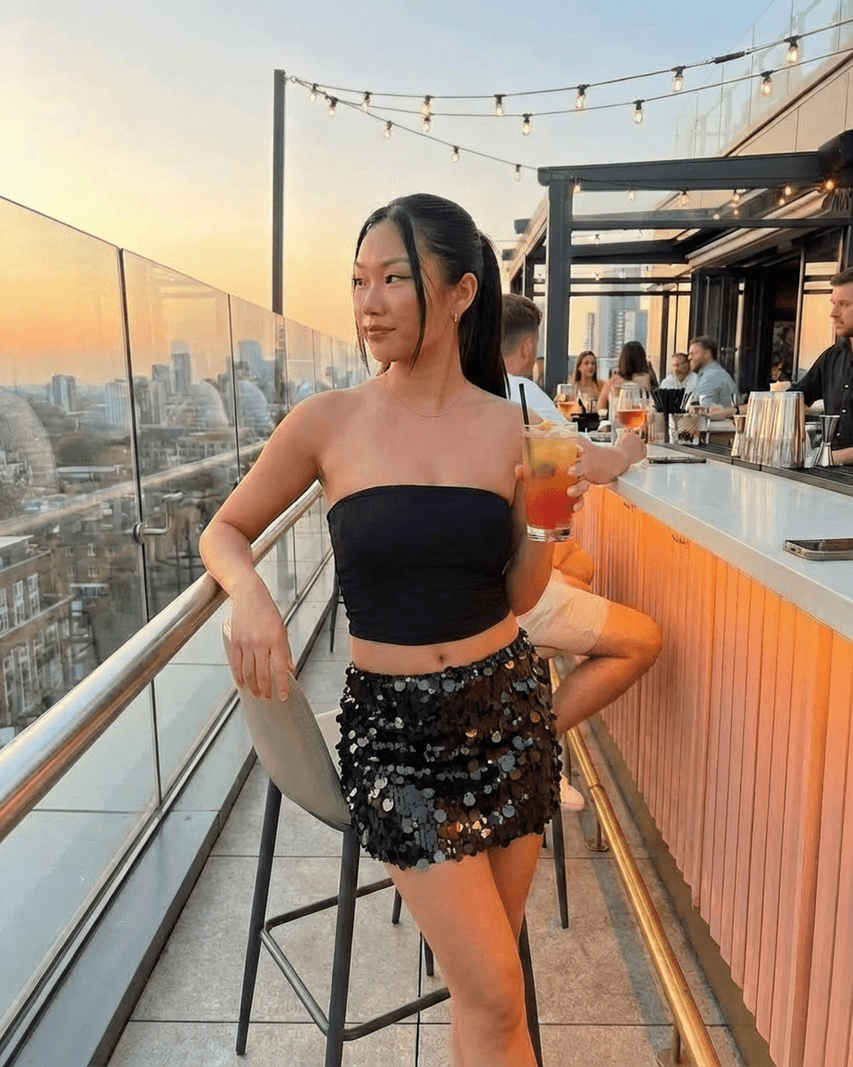 Noir Mini Skirt worn by a stylish woman in a chic rooftop setting, showcasing its black sequin design and A-line silhouette.