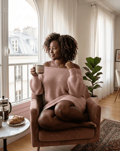 Woman in a soft pink ribbed knit off-the-shoulder dress enjoying coffee, featuring the Nova Mini Dress style by SLIMONA.