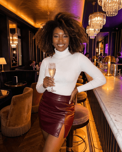 Woman in a white turtleneck holding champagne wearing a Bordeaux Mini Skirt by SLIMONA with a front slit and A-line silhouette.