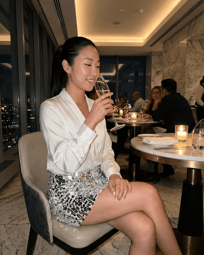 Woman enjoying a drink wearing the Celeste Mini Skirt by SLIMONA in a glamorous dining setting.