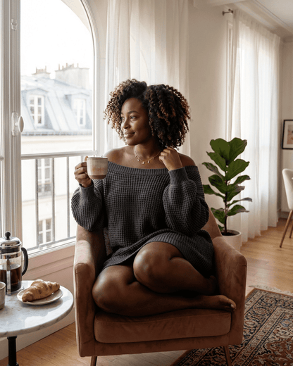 Woman in Ember Mini Dress by SLIMONA, relaxing in a cozy chair with a coffee, showcasing a stylish off-the-shoulder design.