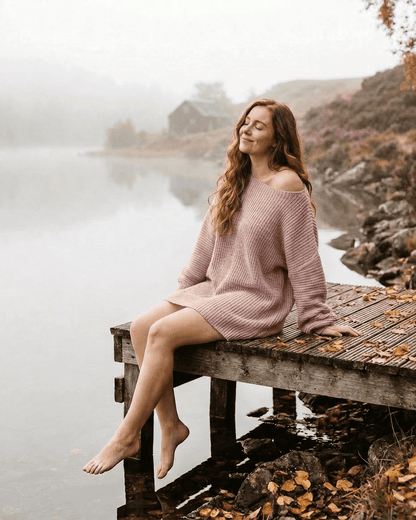 Model wearing a SLIMONA Nova Mini Dress, a soft pink ribbed knit off-the-shoulder, sitting by a tranquil lake.