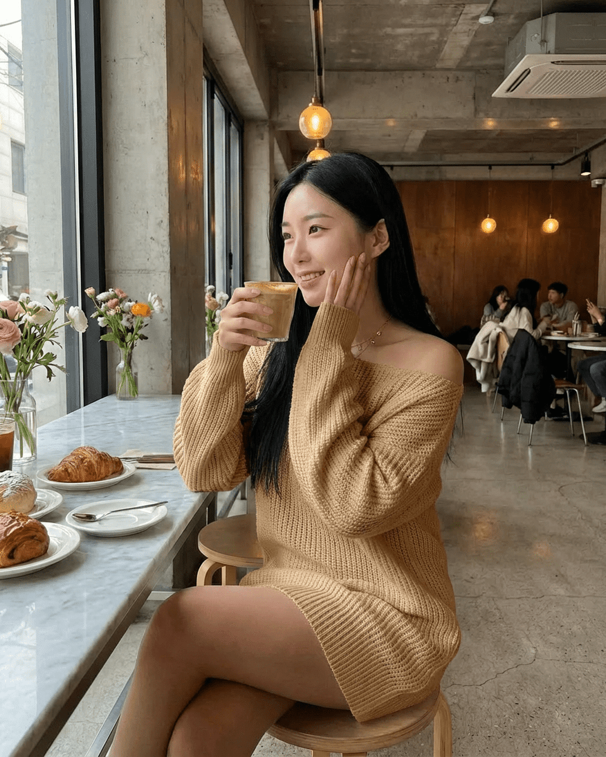 Woman enjoying coffee in cozy off-the-shoulder knit sweater at a café, surrounded by flowers and pastries.