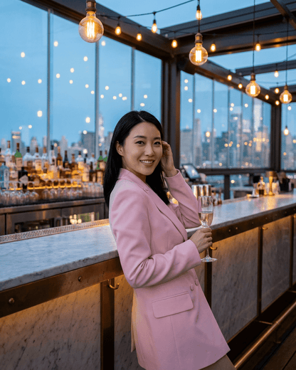 A woman wearing a Rosalie Blazer by SLIMONA, holding a drink at a stylish bar with city lights in the background.