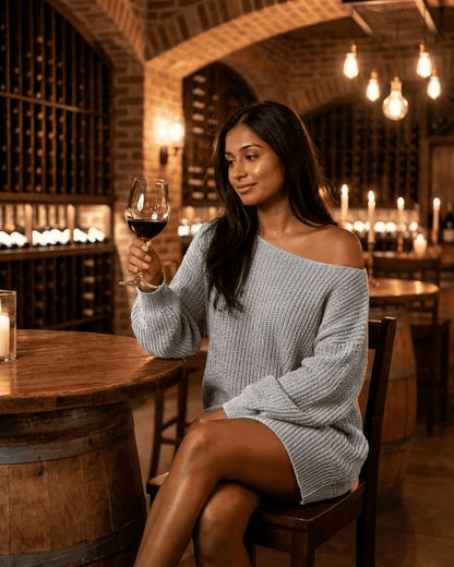Woman enjoying a glass of wine wearing a cozy grey Selene Mini Dress in a warm, rustic wine bar setting.