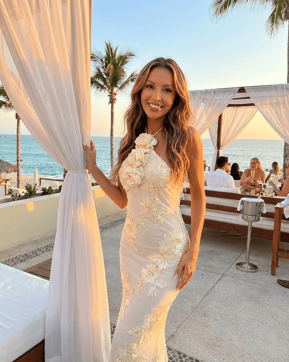 A woman wearing the Valeria Maxi Dress by SLIMONA, showcasing a halter neck and 3D floral foil print against a beach backdrop.