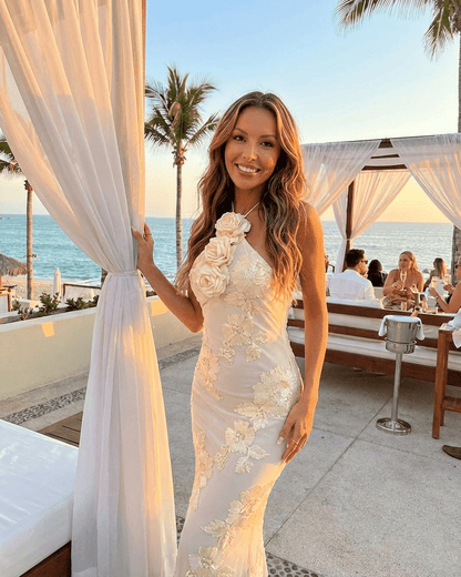 A woman wearing the Valeria Maxi Dress by SLIMONA, showcasing a halter neck and 3D floral foil print against a beach backdrop.