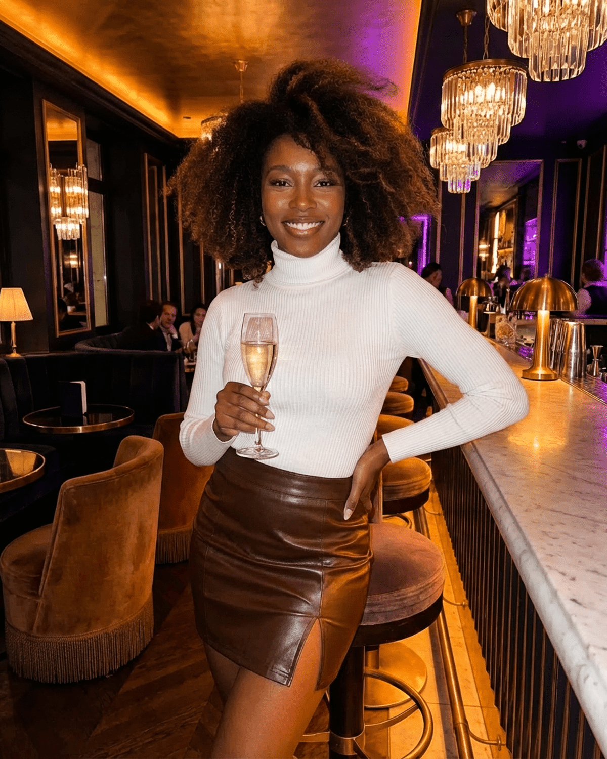 Woman in a stylish outfit featuring the Umbria Mini Skirt by SLIMONA, smiling with a glass of champagne in a chic bar.