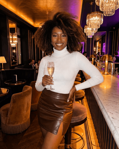 Woman in a stylish outfit featuring the Umbria Mini Skirt by SLIMONA, smiling with a glass of champagne in a chic bar.