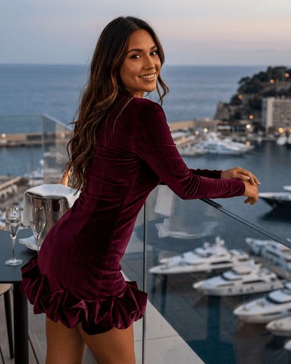Woman wearing Velora Mini Dress by SLIMONA with velvet texture and pleated hem, overlooking a marina at sunset.