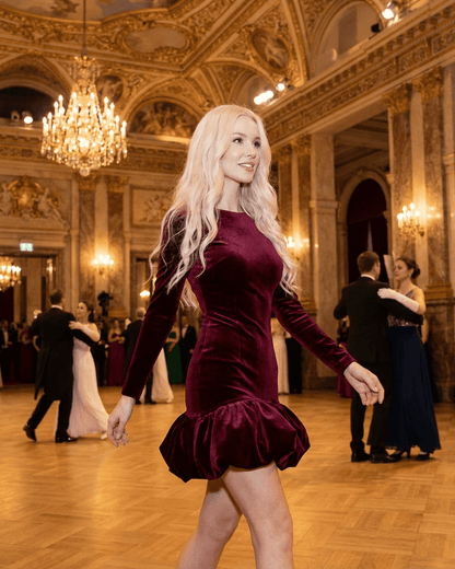 Stylish woman wearing Velora Mini Dress by SLIMONA, showcasing luxe velvet and a pleated hem in a grand ballroom setting.